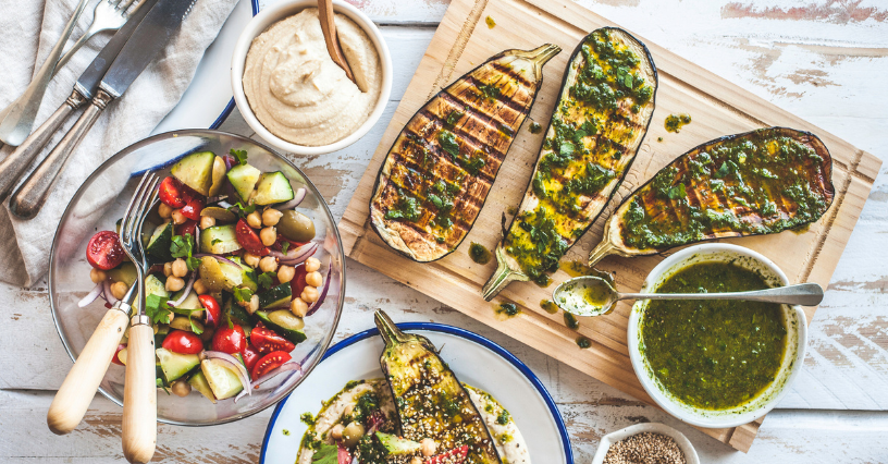 Na desce leżą grillowane bakłażany z ziołowym sosem, obok sałatka i hummus
