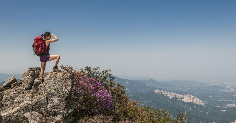 Kobieta w butach trekkingowych, odzieży i plecaku turystycznym obserwująca górski krajobraz ze skały